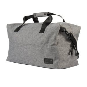 A gray duffel bag with black handles, featuring a side zipper and buckle closure. The bag also displays a small black patch with text near the bottom corner.