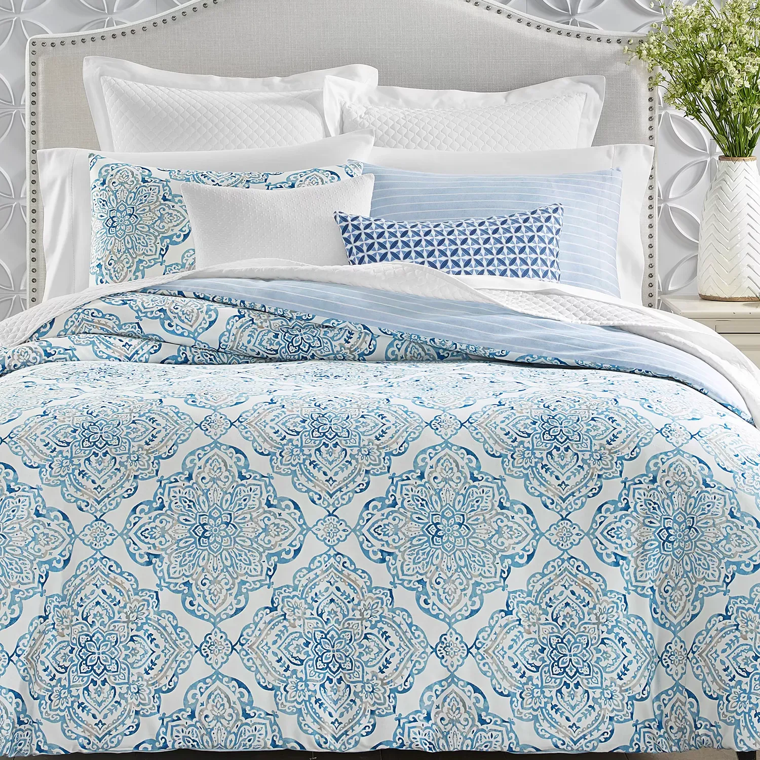 A bed features a patterned blue and white comforter with matching shams, complemented by various textured and printed pillows. The headboard is light gray with a studded border, set against a wall with raised circular designs.