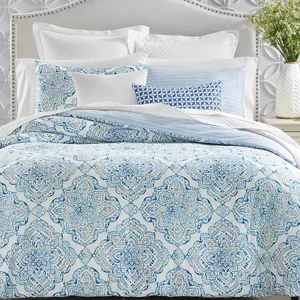A bed features a patterned blue and white comforter with matching shams, complemented by various textured and printed pillows. The headboard is light gray with a studded border, set against a wall with raised circular designs.