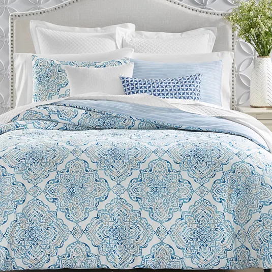 A bed features a patterned blue and white comforter with matching shams, complemented by various textured and printed pillows. The headboard is light gray with a studded border, set against a wall with raised circular designs.