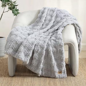 A plush UGG faux-fur throw with a textured stripe pattern is draped over a white chair, presented in a cozy home setting.