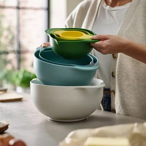 A set of nesting kitchen bowls in various colors, including a strainer and measuring cups, being stacked together on a countertop.