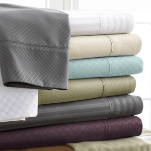 A stack of neatly folded bed sheets in various colors and patterns, including gray, white, beige, teal, olive green, and dark purple, is displayed on a wooden surface.