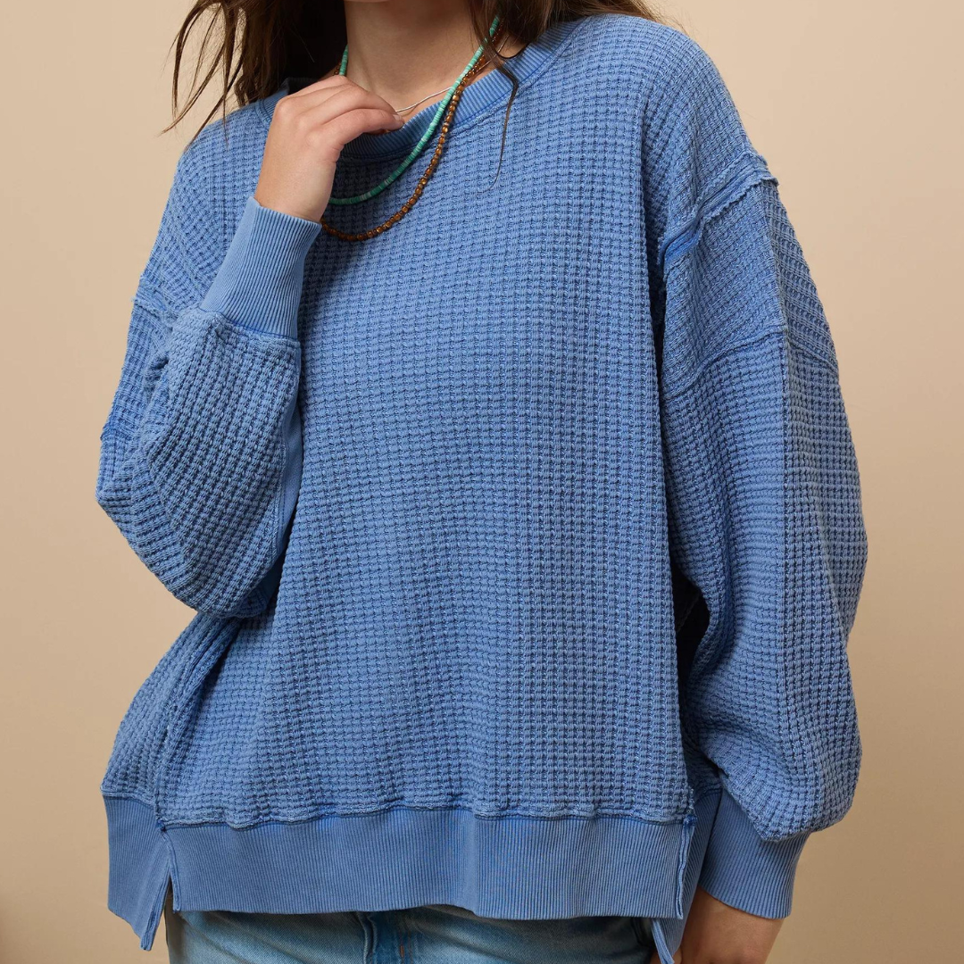 A person is wearing a blue waffle knit sweater with a ribbed neckline and cuffs, paired with a multicolor beaded necklace.