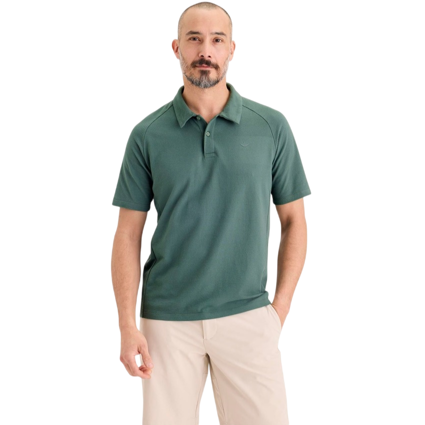 A man is wearing a green polo shirt with beige shorts, standing against a neutral background.