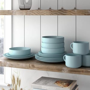 Light blue dinnerware set with plates, bowls, and mugs is displayed on a wooden shelf alongside wine glasses and two books, including \“Flour Water Salt Yeast.\“