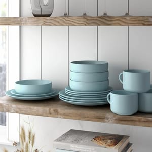 Light blue dinnerware set with plates, bowls, and mugs is displayed on a wooden shelf alongside wine glasses and two books, including \“Flour Water Salt Yeast.\“