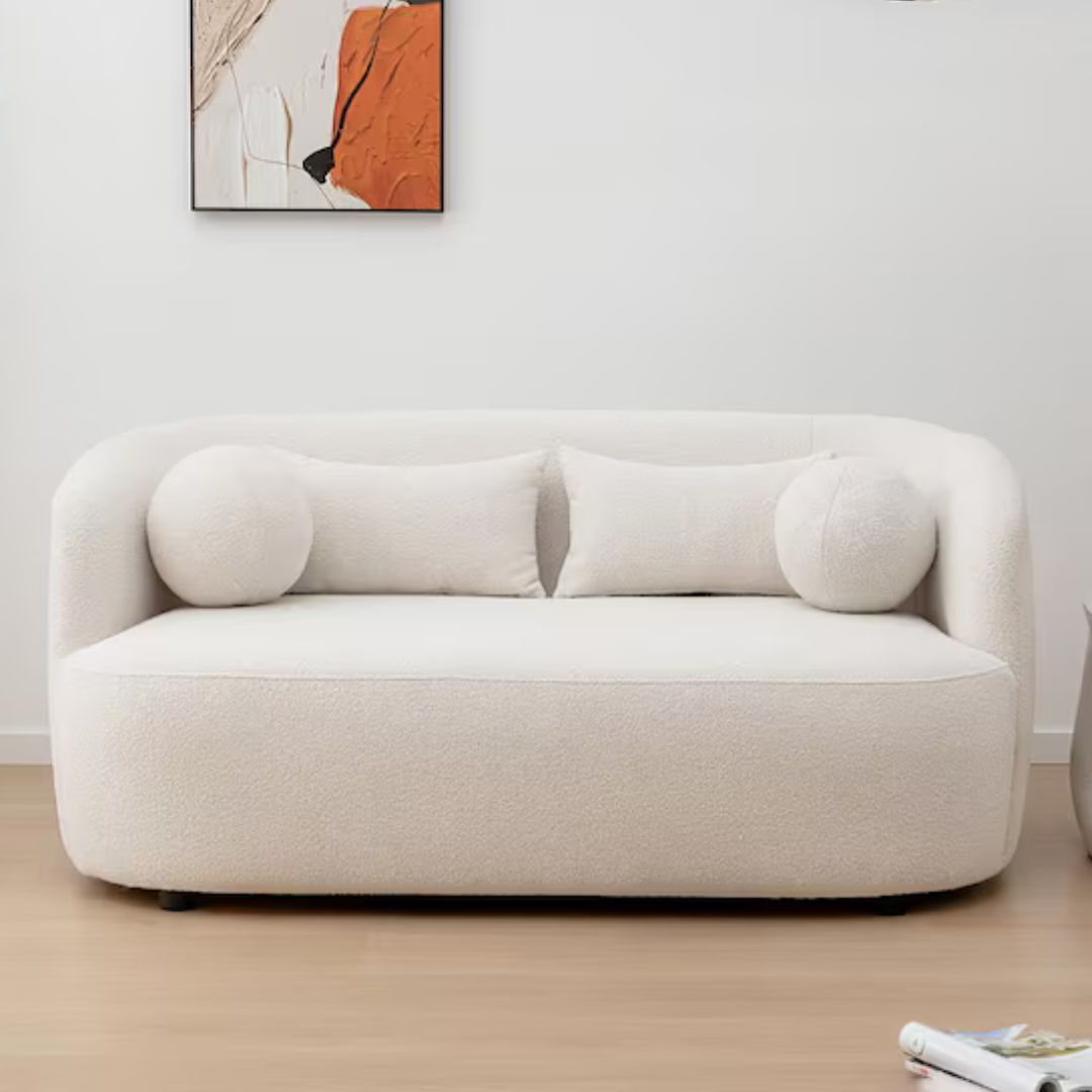 A plush, white loveseat with rounded edges includes two round bolster pillows and a rectangular cushion on a wooden floor, beneath an abstract wall art piece in tones of orange, black, and white.