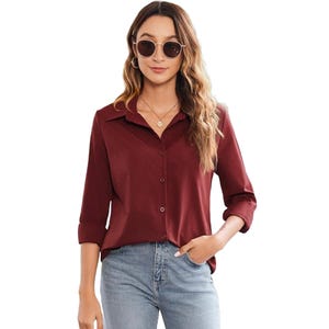 A woman is wearing a long-sleeve, maroon button-down blouse paired with light blue jeans. She accessorizes with round sunglasses, a necklace, and has her hair loose, styled with waves.