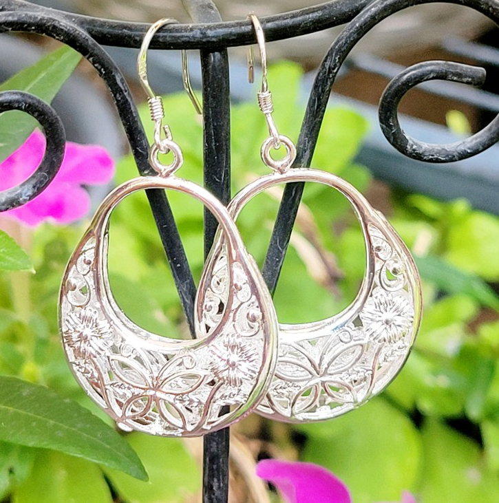 Intricately designed metal earrings featuring floral and geometric patterns, hanging from a decorative display stand with a backdrop of flowers.