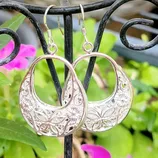 Intricately designed metal earrings featuring floral and geometric patterns, hanging from a decorative display stand with a backdrop of flowers.