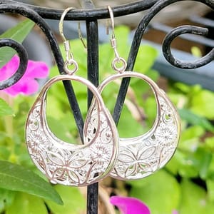Intricately designed metal earrings featuring floral and geometric patterns, hanging from a decorative display stand with a backdrop of flowers.