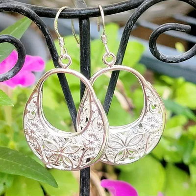 Intricately designed metal earrings featuring floral and geometric patterns, hanging from a decorative display stand with a backdrop of flowers.