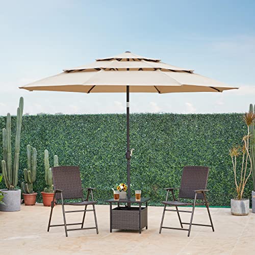 A large beige multi-tier patio umbrella is featured, providing shade over a table with two brown woven chairs. A green hedge serves as a backdrop.