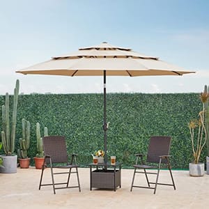 A large beige multi-tier patio umbrella is featured, providing shade over a table with two brown woven chairs. A green hedge serves as a backdrop.