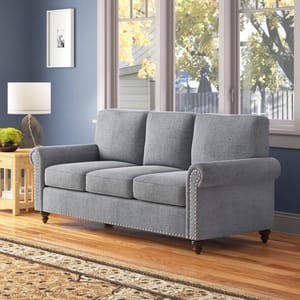 Gray upholstered sofa with rolled arms and nailhead trim on a wooden floor against a blue wall.