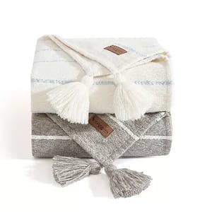 Two stacked Koolaburra by UGG woven throws with tassel accents, one in white with blue stripes and the other in gray.