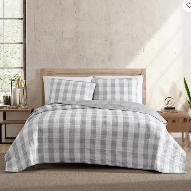 A neatly made bed with a gray and white plaid bedding set, including a quilt and matching pillow shams. The room features a natural wood headboard, a potted plant, a small desk with decor items, and a suitcase-style nightstand.