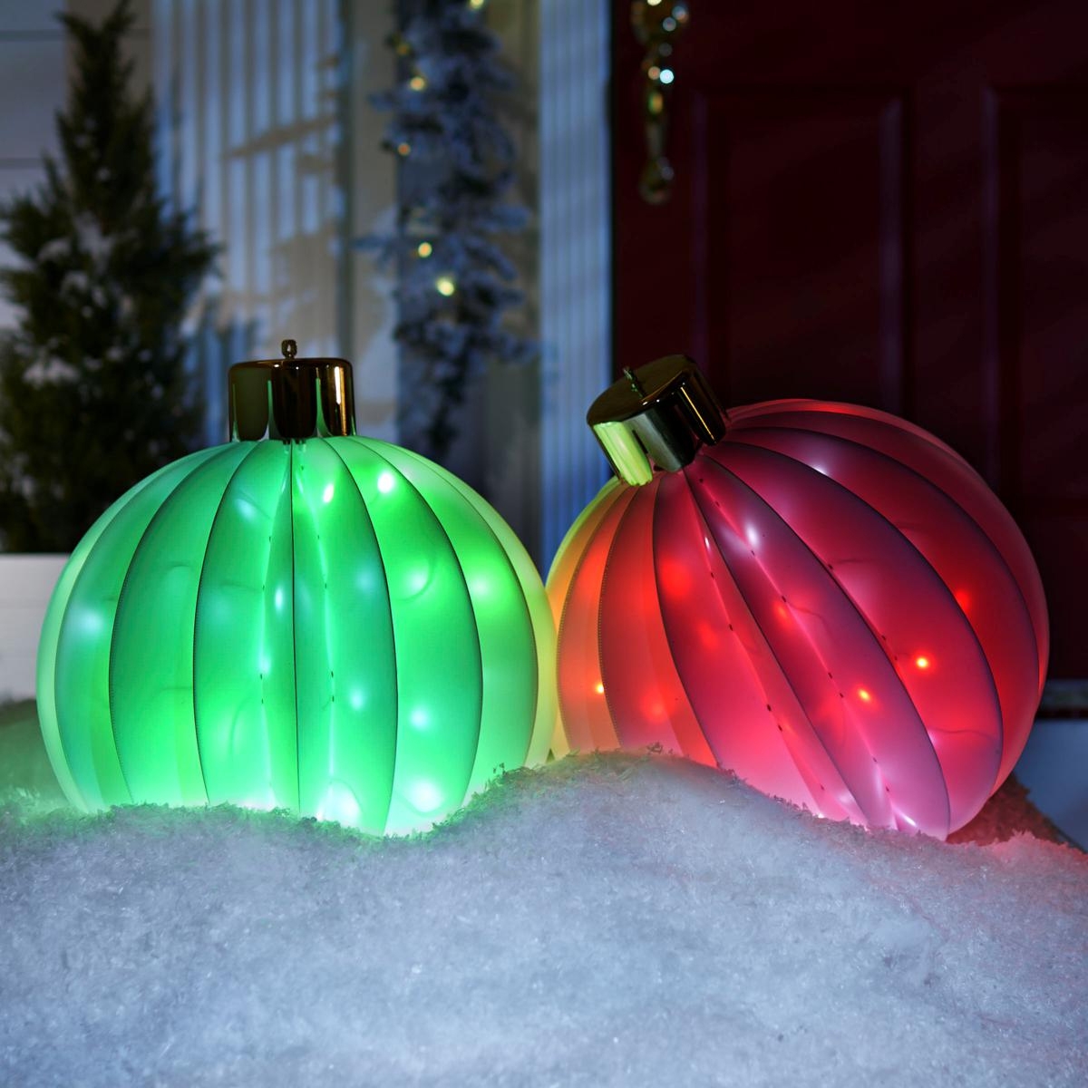 Two brightly illuminated foldable holiday orbs, one green and one red, rest on a snowy surface with a festive backdrop.