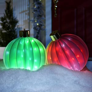 Two brightly illuminated foldable holiday orbs, one green and one red, rest on a snowy surface with a festive backdrop.