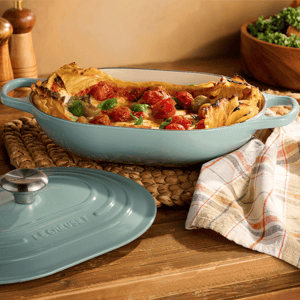 A light blue oval Le Creuset casserole dish is on a wooden table, partially covered with a plaid cloth, containing a baked dish with layered pasta, cherry tomatoes, and herbs. The lid is nearby, and there are salt and pepper shakers in the background.