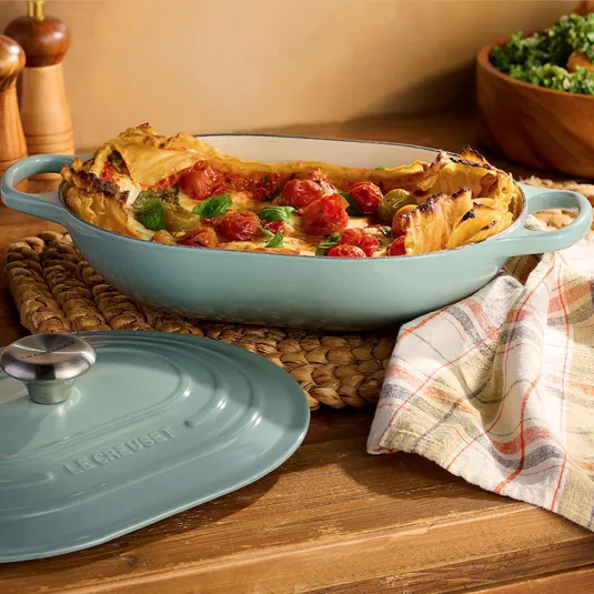 A light blue oval Le Creuset casserole dish is on a wooden table, partially covered with a plaid cloth, containing a baked dish with layered pasta, cherry tomatoes, and herbs. The lid is nearby, and there are salt and pepper shakers in the background.