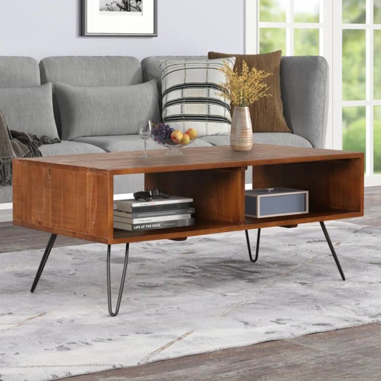 The Trent Austin Jorgenson Coffee Table features a wooden rectangular design with open shelving and black metal hairpin legs. It's styled in a modern living room setting with decor items like books and a vase.
