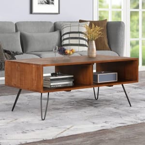 The Trent Austin Jorgenson Coffee Table features a wooden rectangular design with open shelving and black metal hairpin legs. It's styled in a modern living room setting with decor items like books and a vase.