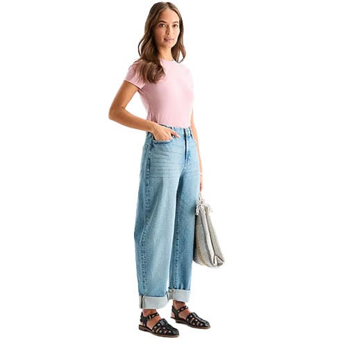A person wears a pink T-shirt, high-waisted, light-wash jeans rolled at the ankles, black cutout sandals, and carries a gray textured shoulder bag.