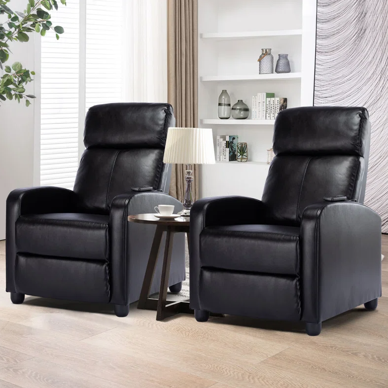Two black leather reclining chairs flank a small round table with a lamp and a cup. The background features shelves with books and decorative items.