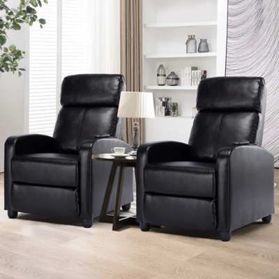 Two black leather reclining chairs flank a small round table with a lamp and a cup. The background features shelves with books and decorative items.