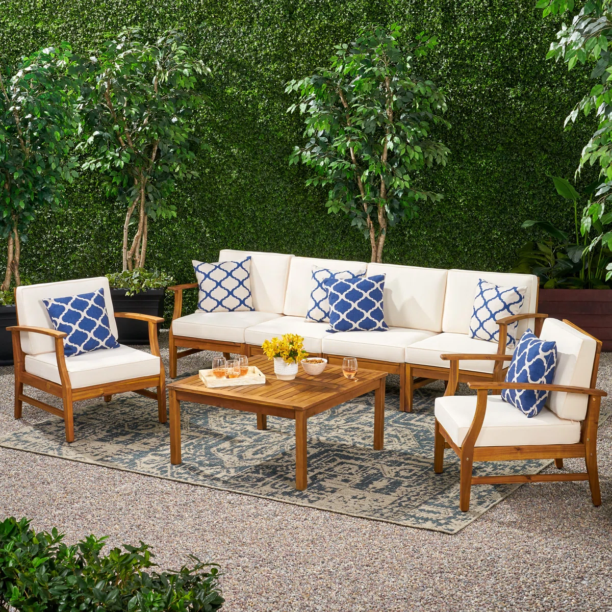 Outdoor patio set featuring a wooden sectional sofa, two armchairs with cream cushions, blue patterned pillows, and a wooden coffee table on a patterned rug, surrounded by lush greenery.