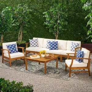 Outdoor patio set featuring a wooden sectional sofa, two armchairs with cream cushions, blue patterned pillows, and a wooden coffee table on a patterned rug, surrounded by lush greenery.
