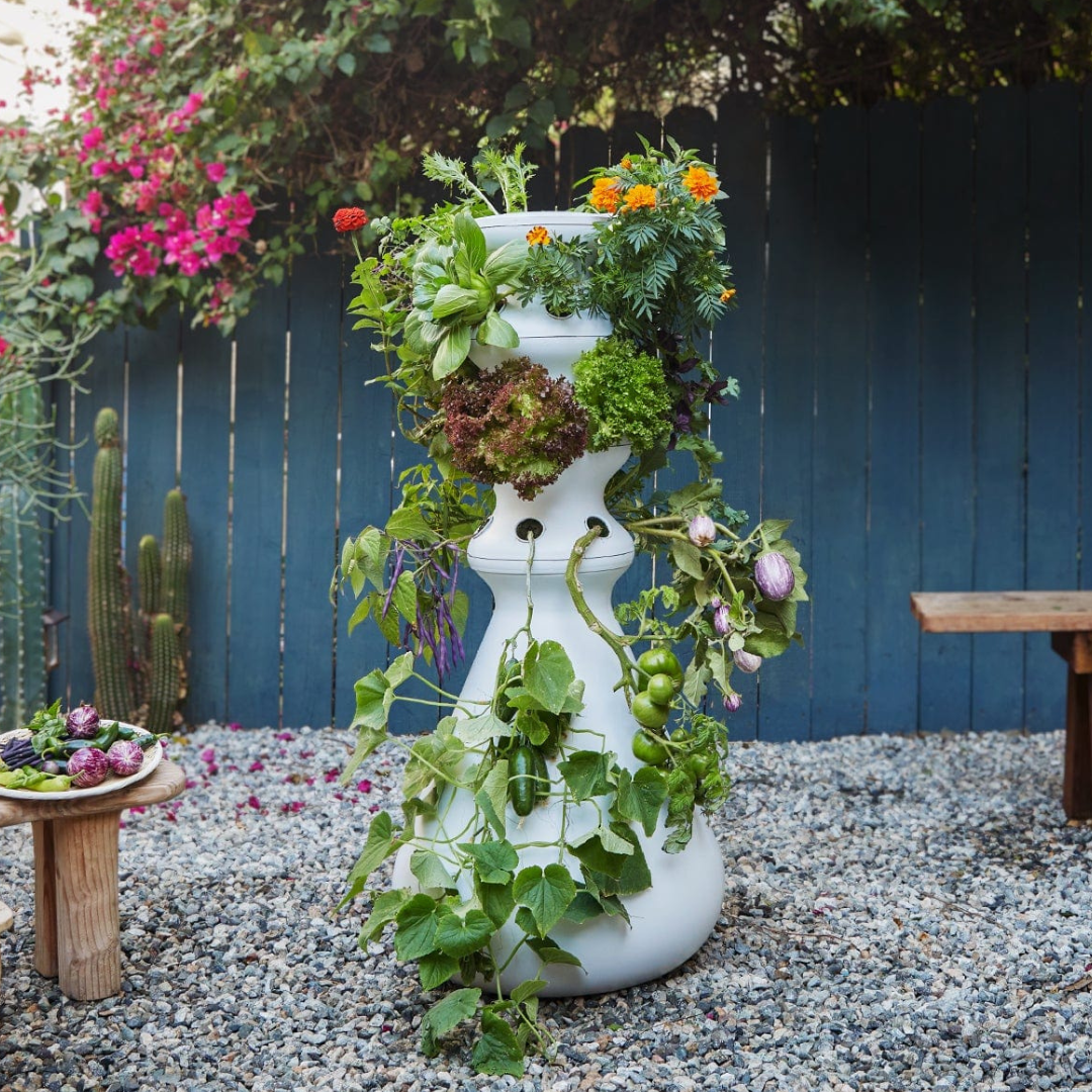 A white tower garden with various plants and vegetables growing from multiple openings, set in a backyard with gravel ground and colorful flowers in the background.