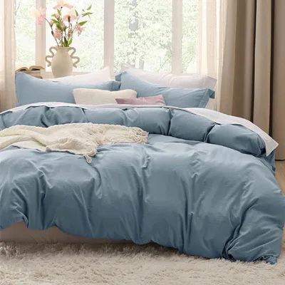A neatly arranged bed with a light blue duvet cover, matching pillows, and a white knit throw blanket on a fluffy rug, with decorative pillows and a vase of flowers nearby.