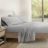 A neatly arranged bedroom with light gray bedding on a wooden bed, next to a bedside table holding a lamp and books. A large window shows a view of trees outside.