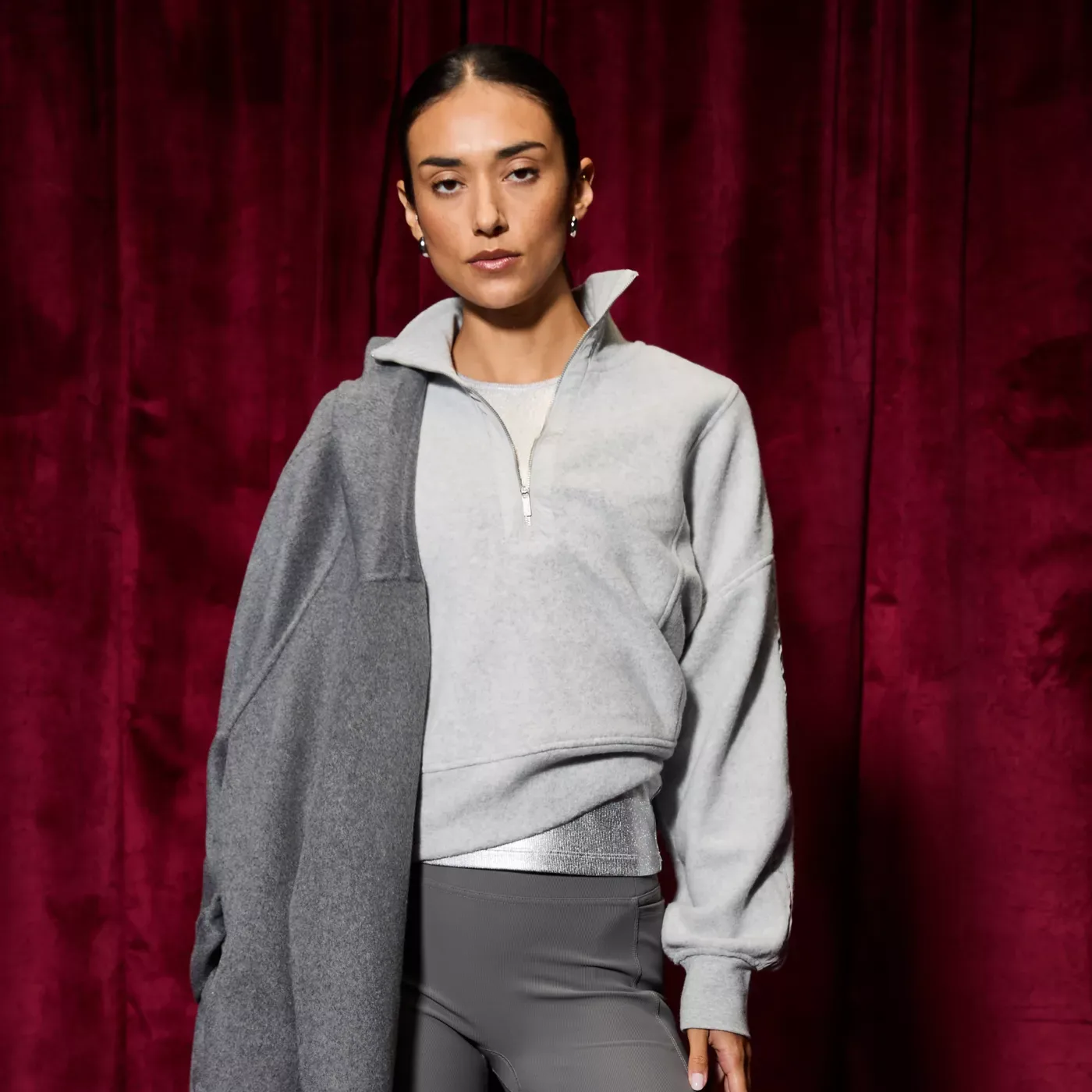 A person wears a light gray half-zip pullover, gray leggings, and holds a gray coat, set against a maroon curtain backdrop.
