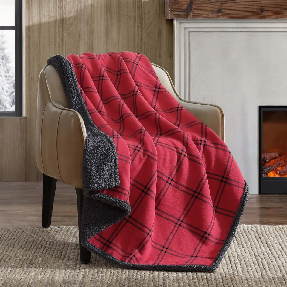 A red plaid blanket with black trim is draped over a beige armchair beside a fireplace in a cozy, snow-covered room.