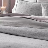 A bed is neatly made with gray bedding, including soft textured comforters, pillows, and a decorative throw. A lit candle sits on a marble surface nearby.