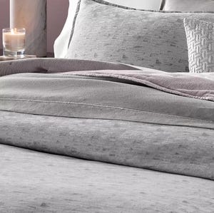 A bed is neatly made with gray bedding, including soft textured comforters, pillows, and a decorative throw. A lit candle sits on a marble surface nearby.