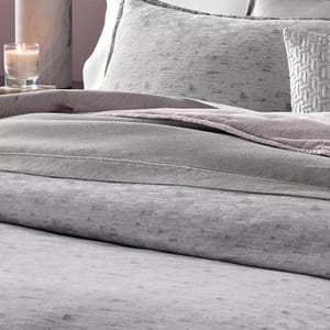 A bed is neatly made with gray bedding, including soft textured comforters, pillows, and a decorative throw. A lit candle sits on a marble surface nearby.