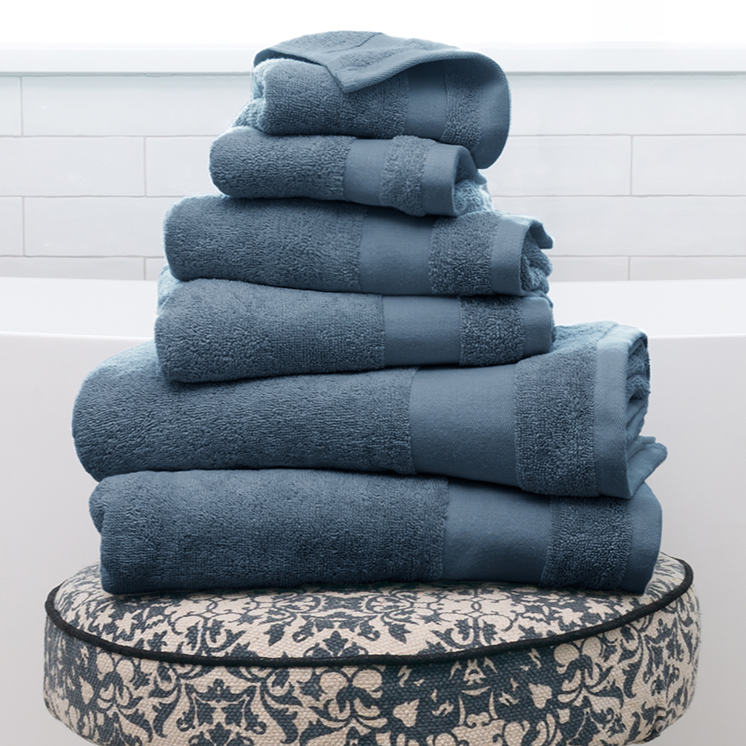 Stack of blue towels on a patterned stool with wooden legs, set in a bright, tiled bathroom.