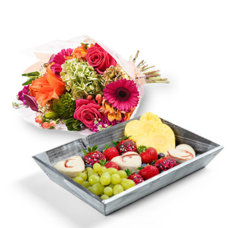 A vibrant bouquet of mixed flowers includes roses, gerbera daisies, and hydrangeas in red, pink, and orange hues. Paired with a dessert tray featuring green grapes, pineapple slices, chocolate-dipped strawberries, and other mixed fruits.
