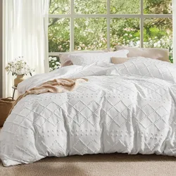 A white quilted bedding set with a textured diamond pattern is displayed in a bright room with large windows overlooking a garden. A beige throw and a vase with flowers are placed beside the bed.