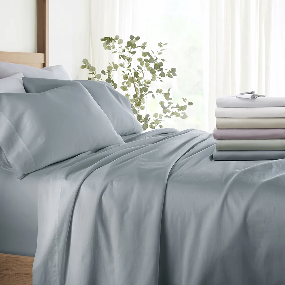 A neatly made bed features soft, light blue 100% cotton sateen sheets, accompanied by a stack of folded sheets in various colors like white, beige, and lavender. A leafy plant and soft curtains create a serene atmosphere.