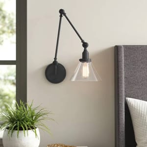 Wall-mounted swing arm sconce with a black metal arm and round base. It features a clear conical glass shade, exposing a visible light bulb, positioned above a grey upholstered headboard.