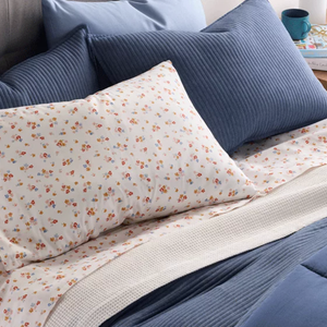 A bed setup featuring blue textured pillows and a white pillow with a small floral print. The bedding includes a white waffle blanket and a blue quilt. A small wooden nightstand with a teal mug and stacked books is visible in the background.