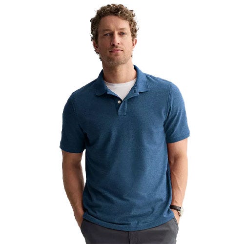 A man in a blue short-sleeve polo shirt and gray pants stands with hands in pockets.