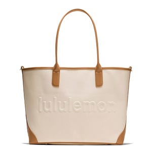 A stylish cream tote bag with tan leather accents, featuring the \“lululemon\“ logo embossed on the front.