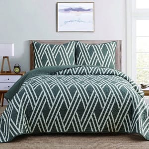 A bed features a geometric-patterned quilt and matching pillows in green and white tones, set against a neutral room with a framed wall art piece above it.